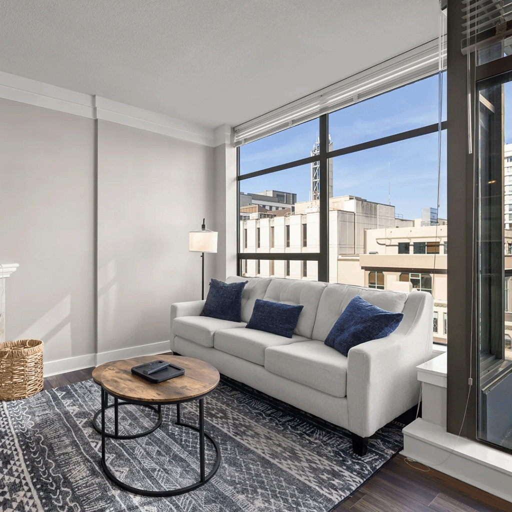 Modern minimalist living room with a white sofa, large windows providing ample sunlight, and a serene view of the urban exterior