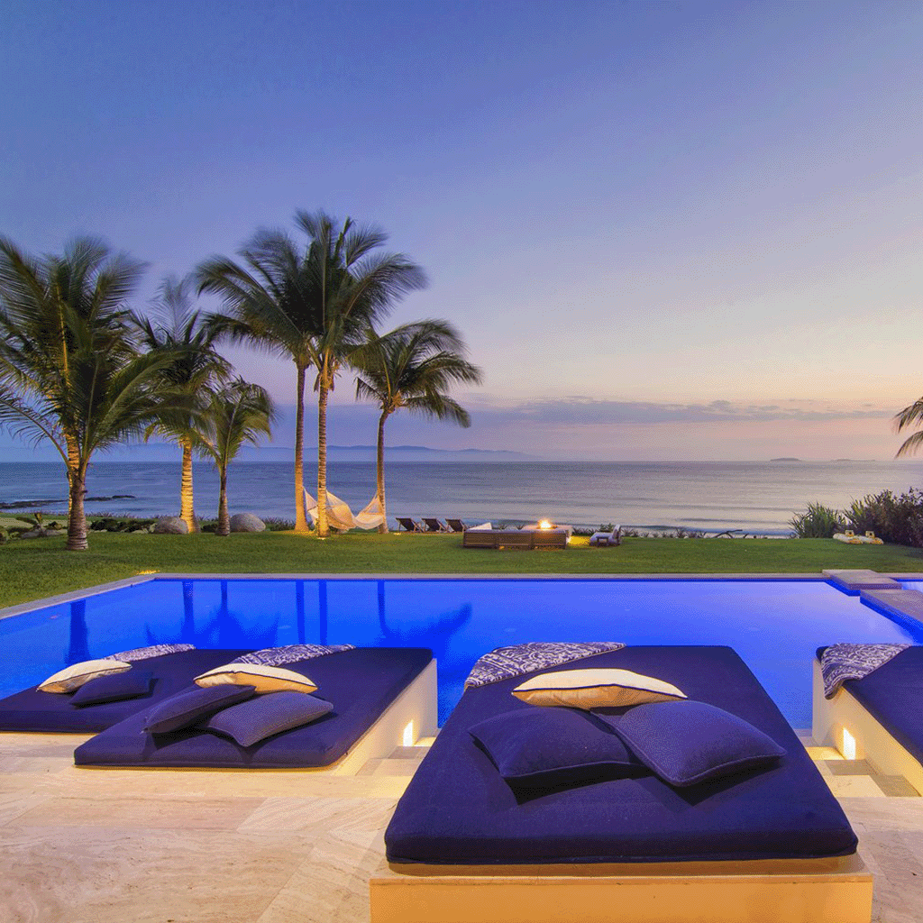 Luxurious poolside lounge area, managed to perfection, with plush seating and palm trees overlooking a calm ocean at dusk, highlighting serene property management.