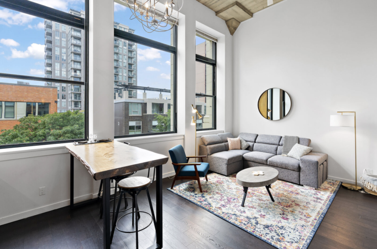 Modern loft with a gray sofa, rustic wooden table, and wide windows offering an urban view