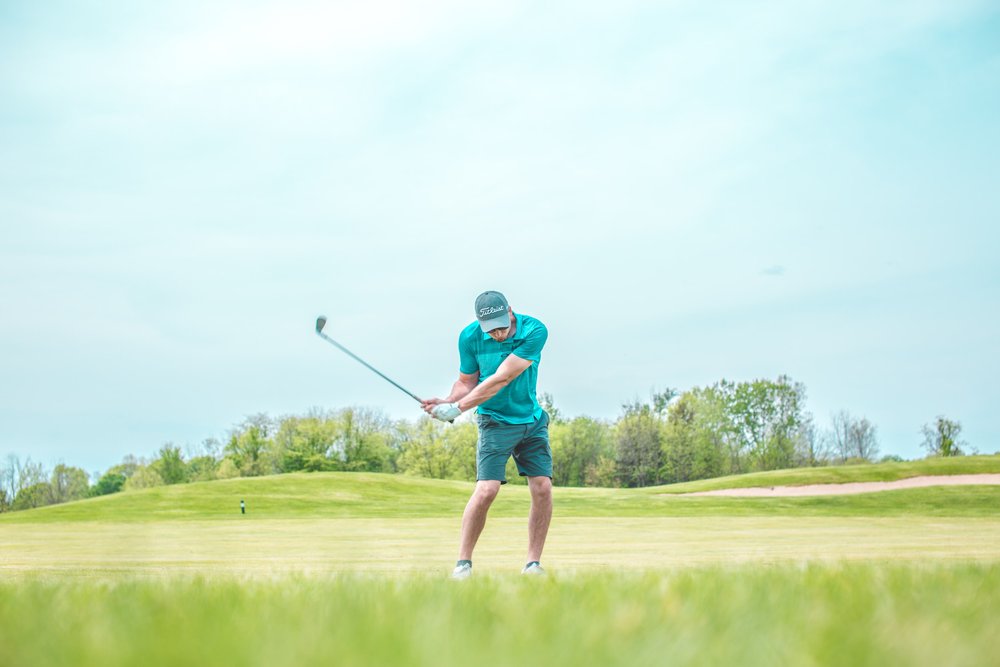 A man in a teal shirt swinging a golf club on a sunny day with a clear blue sky and lush green course.