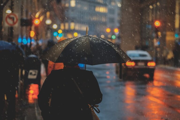 A lone figure holding an umbrella, illuminated by city lights reflecting on wet streets during a nighttime rain.